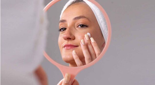 Mulher com faixa no cabelo aplicando creme no rosto enquanto se observa no espelho de mão.