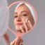 Mulher com faixa no cabelo aplicando creme no rosto enquanto se observa no espelho de mão.
