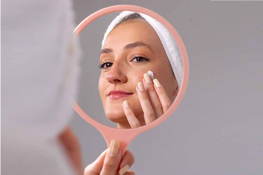 Mulher com faixa no cabelo aplicando creme no rosto enquanto se observa no espelho de mão.