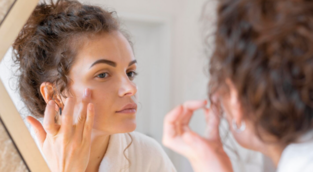 Mulher aplica produto de skincare no rosto enquanto se observa no espelho durante a rotina de cuidados com a pele.