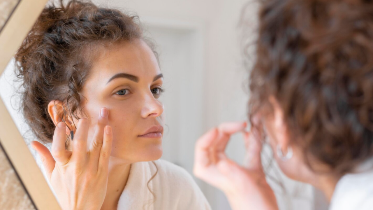 Mulher aplica produto de skincare no rosto enquanto se observa no espelho durante a rotina de cuidados com a pele.