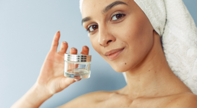 Mulher com toalha na cabeça segura um pote de creme para cuidados com a pele e sorri.