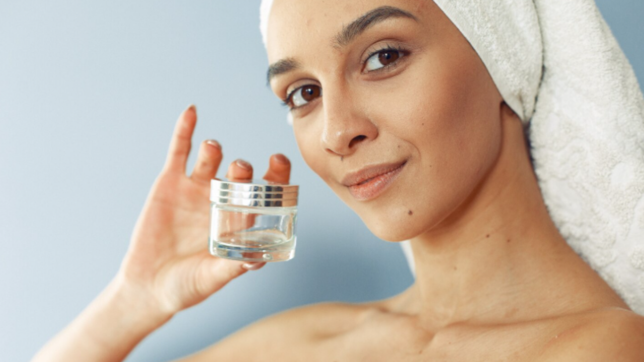 Mulher com toalha na cabeça segura um pote de creme para cuidados com a pele e sorri.