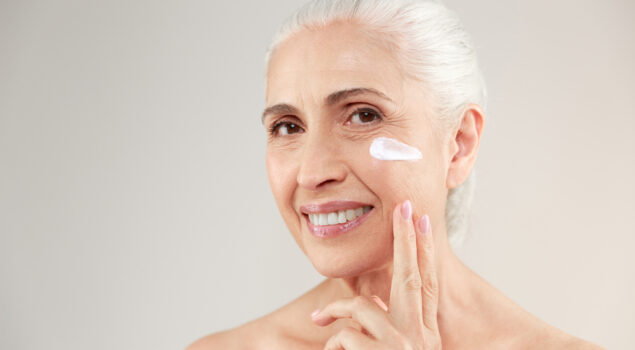 Mulher de cabelos brancos sorri para a câmera enquanto passa um produto de pele na bochecha, representando a importância da skincare personalizada.