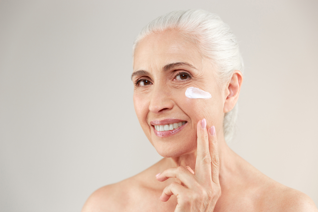 Mulher de cabelos brancos sorri para a câmera enquanto passa um produto de pele na bochecha, representando a importância da skincare personalizada.